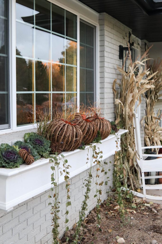A picture of wicker pumpkins and flowering kale in a white window box.